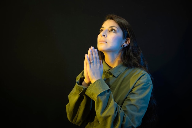 Mujer joven en uniforme militar rezando por Ucrania sobre un fondo negro con iluminación de estudio