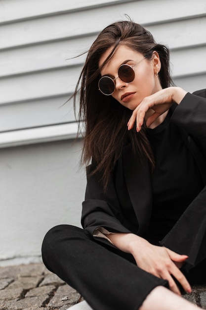 La mujer joven urbana sexy de moda en gafas de sol redondas de moda en una  elegante chaqueta negra para jóvenes disfruta de un descanso junto a la  pared de madera vintage