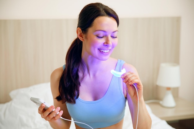 Una mujer joven usa un complejo de blanqueamiento para blanquear los dientes en el hogar lámpara usb uv para blanquear los dientes