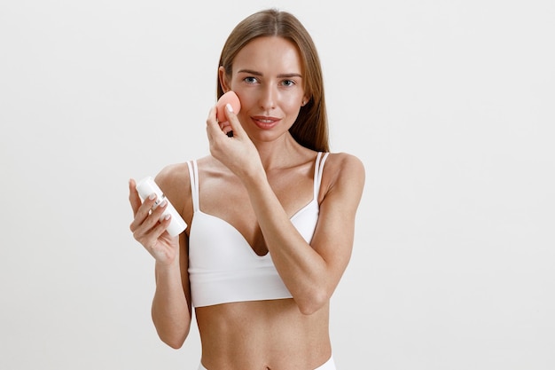 Mujer joven usa esponja rosa para aplicar productos cosméticos de belleza en la cara en el fondo del estudio