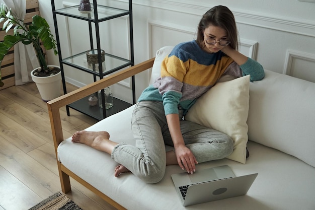 Mujer joven usando una computadora portátil escribiendo usando un panel táctil sentado en casa en un sofá trabajo remoto en casa