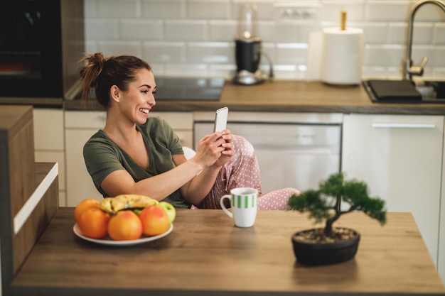 Mujer joven usando su teléfono inteligente mientras toma café por la mañana en casa.