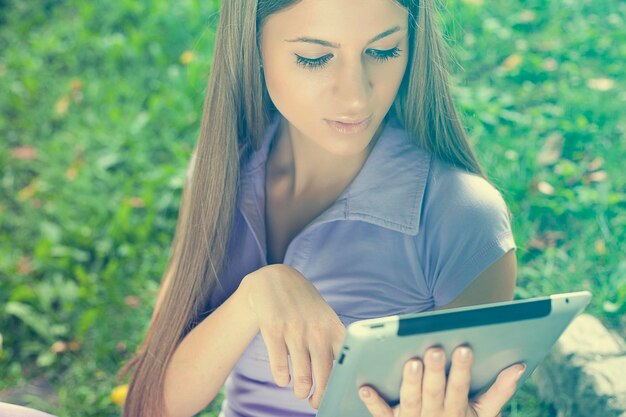 Foto mujer joven usando una tableta digital mientras está sentada en un campo de hierba en el parque