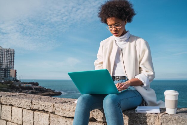 Mujer joven usando el teléfono mientras está sentada junto al mar contra el cielo