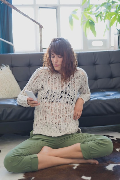 Foto mujer joven usando el teléfono mientras está sentada contra el sofá en casa