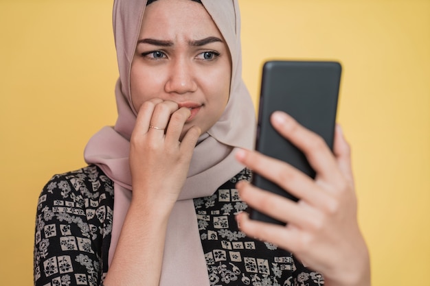 Mujer joven con velo sorprendido y preocupado mientras mira la pantalla del teléfono móvil con gesto de dedo mordido