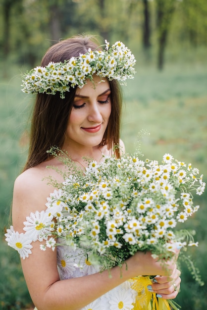 Una mujer joven vestida con un hermoso vestido tierno y una corona sostiene un ramo de manzanilla en sus manos