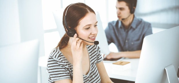 Mujer joven vestida de manera informal usando auriculares y computadora mientras habla con los clientes en línea