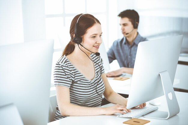 Foto mujer joven vestida de manera informal usando auriculares y computadora mientras habla con los clientes en línea