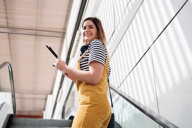 Mujer joven vestida con monos amarillos subiendo las escaleras mecánicas usando la tecnología de estilo de vida Concept de la tableta al aire libre