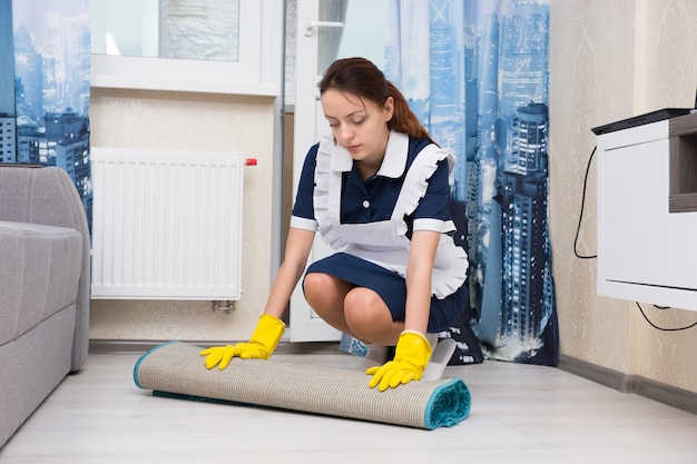 Mujer joven vestida con un uniforme pulcro y delantal blanco haciendo las tareas de limpieza agachándose para enrollar una alfombra en una sala de estar para limpiar el piso debajo