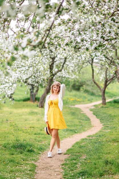 Una mujer joven con un vestido amarillo y un cardigan blanco camina en un jardín de flores de primavera.