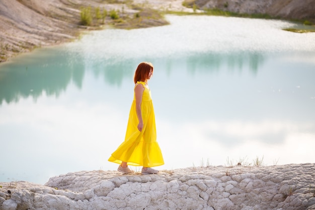 Mujer joven con un vestido amarillo cerca del lago con agua azul y árboles verdes.