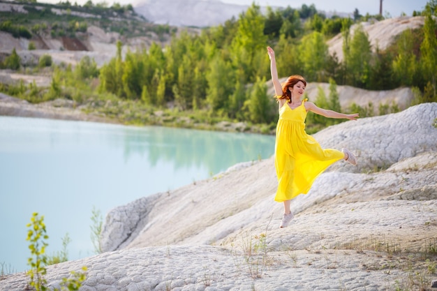 Mujer joven con un vestido amarillo cerca del lago con agua azul y árboles verdes.