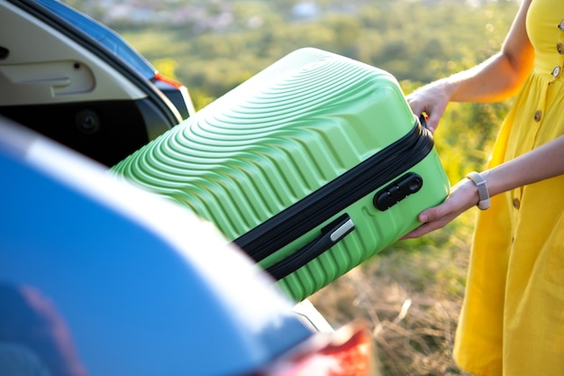 Mujer joven en vestido amarillo de verano tomando la maleta verde del maletero del coche. Concepto de viajes y vacaciones.