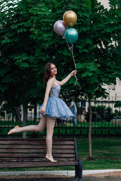 Una mujer joven con un vestido azul camina descalza sobre un banco de madera y sostiene globos voladores fingiendo ...