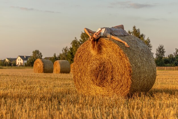 Mujer joven en vestido azul de cerca en el fondo del campo con un pajar al atardecer.