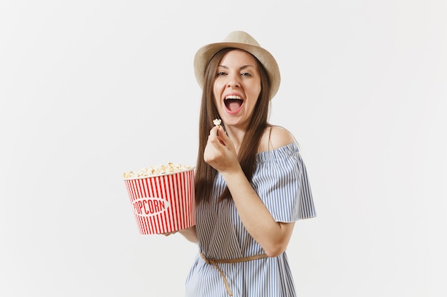 Mujer joven en vestido azul, sombrero viendo la película sosteniendo comiendo palomitas de maíz de cubo aislado sobre fondo blanco. Gente, emociones sinceras en el cine, concepto de estilo de vida. Área de publicidad. Copie el espacio.