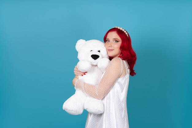 Mujer joven con un vestido blanco con pelo rojo abrazando un oso de peluche blanco