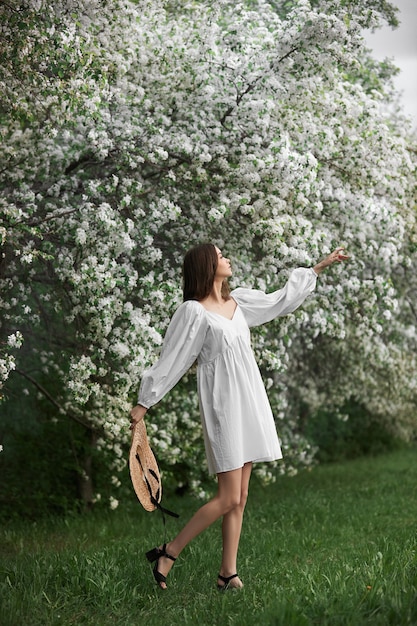 Mujer joven con vestido blanco con sombrero de paja camina por el parque floreciente del jardín de la primavera. Ha llegado la primavera, estado de ánimo romántico