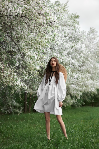 Mujer joven con vestido blanco con sombrero de paja camina por el parque floreciente del jardín de la primavera. Ha llegado la primavera, estado de ánimo romántico