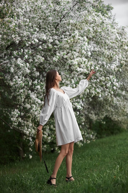 Mujer joven con vestido blanco con sombrero de paja camina por el parque floreciente del jardín de la primavera. Ha llegado la primavera, estado de ánimo romántico