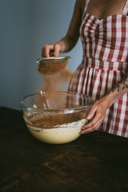 Foto mujer joven con un vestido a cuadros rojo y blanco horneando un pastel, tamizando el cacao en polvo en un vaso grande