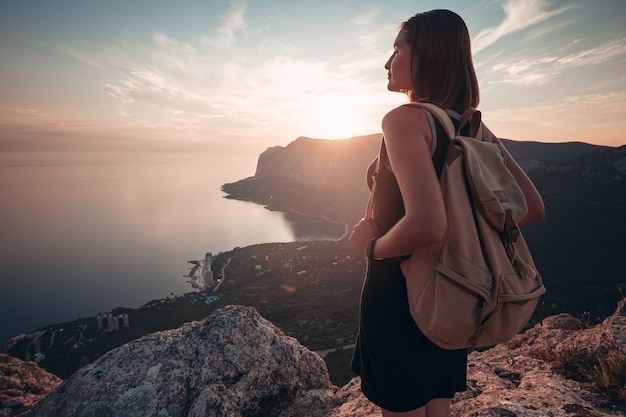 Mujer joven con un vestido deportivo con mochila disfruta de la vista de las montañas y el mar al atardecer. Viajes y concepto de estilo de vida activo. Glamping