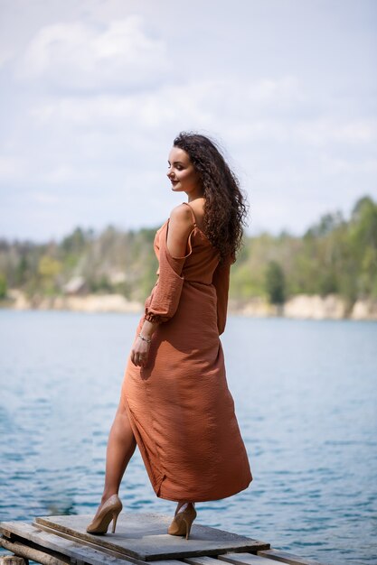 Una mujer joven con un vestido se encuentra en mampostería de madera en medio de un lago azul. Niña feliz sonríe y brilla el sol, día de verano. Tiene el pelo largo y rizado y apariencia europea.