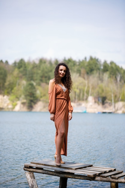 Una mujer joven con un vestido se encuentra en mampostería de madera en medio de un lago azul. Niña feliz sonríe y brilla el sol, día de verano. Tiene el pelo largo y rizado y apariencia europea.