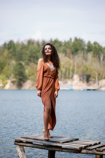 Una mujer joven con un vestido se encuentra en mampostería de madera en medio de un lago azul. Niña feliz sonríe y brilla el sol, día de verano. Tiene el pelo largo y rizado y apariencia europea.