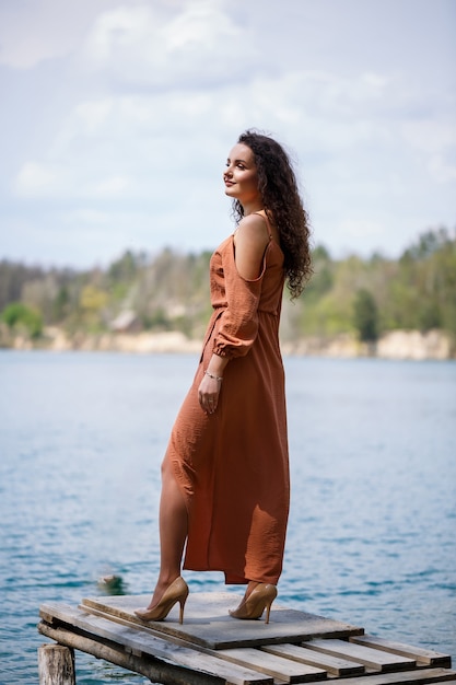 Una mujer joven con un vestido se encuentra en mampostería de madera en medio de un lago azul. Niña feliz sonríe y brilla el sol, día de verano. Tiene el pelo largo y rizado y apariencia europea.