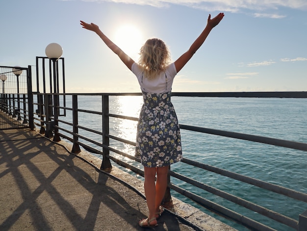 Mujer joven en vestido está de pie con las manos levantadas, en el muelle desierto en el fondo del mar y el cielo