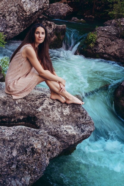 La mujer joven con el vestido está sentada en la piedra en medio de un arroyo Tonificación de moda