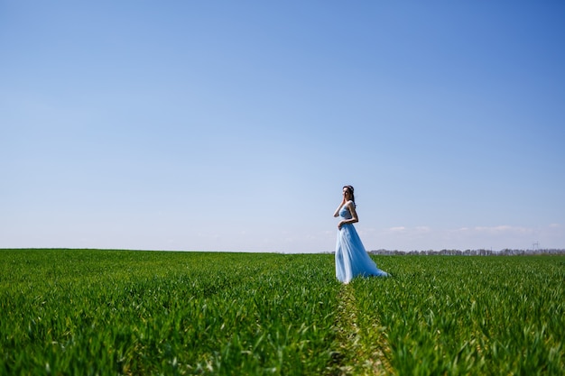 Mujer joven con un vestido largo azul sobre un fondo de campo verde. Retrato de moda de una hermosa niña con una sonrisa en su rostro