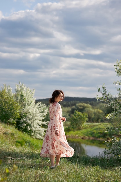 Mujer joven en vestido largo camina en el jardín de primavera cerca del lago. Chica descansa en la naturaleza, ha llegado la primavera.