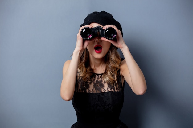 Mujer joven en vestido negro con binoculares en pared gris