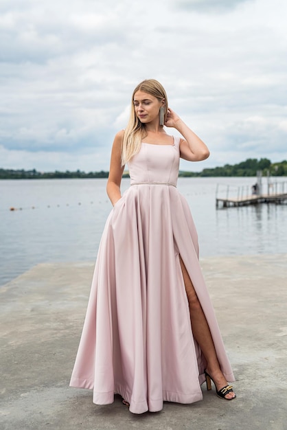 Mujer joven en vestido de noche largo rosa de pie sola cerca del lago en el parque de la ciudad