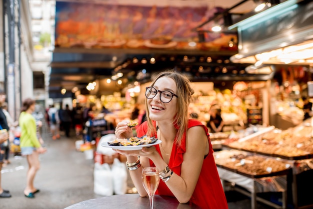 Mujer joven en vestido rojo almorzando con mejillones y vino rosado sentado en el mercado de alimentos