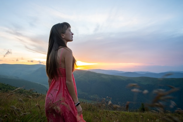 Mujer joven en vestido rojo de pie sobre el césped en una tarde ventosa en las montañas de otoño disfrutando de la vista de la naturaleza.