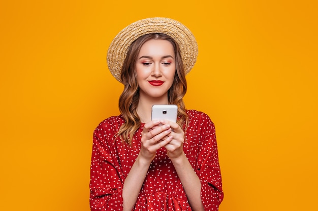 Mujer joven en un vestido rojo de verano sombrero de paja charlando por teléfono móvil aislado sobre la pared de la pared de color naranja. Pedidos en línea compras concepto de crédito comercial.