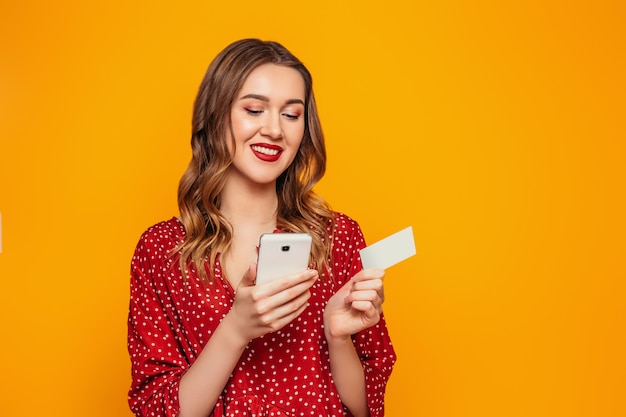 La mujer joven en un vestido rojo del verano sostiene un teléfono móvil y una tarjeta de crédito en sus manos aisladas en una pared anaranjada con la maqueta. Niña mira el teléfono y hace compras en línea