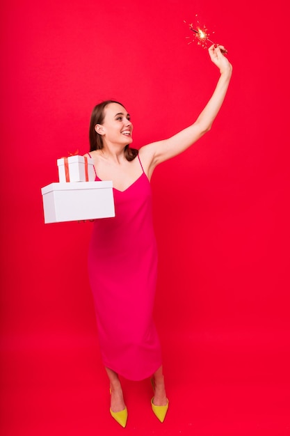 Una mujer joven con un vestido rosa posa con regalos de Navidad en cajas y bengalas en un fondo rojo