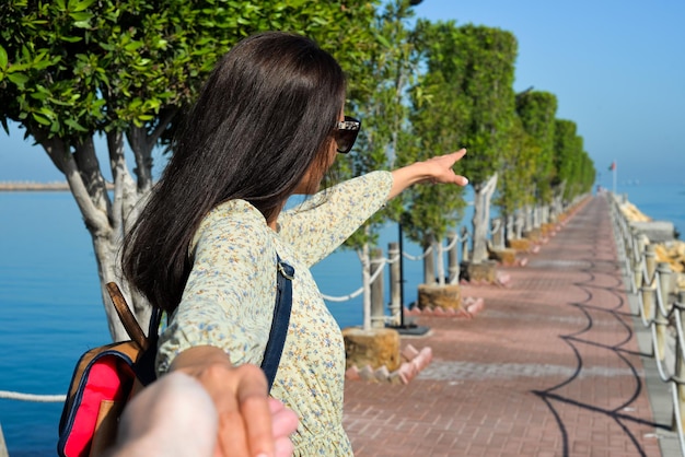 Una mujer joven con un vestido señala la dirección al mar con la mano Pier en la playa de Sharjah