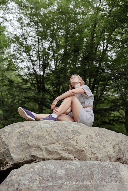 Mujer joven en vestido de verano sentada sobre una piedra grande en el bosque mirando a otro lado