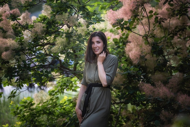 Foto una mujer joven con un vestido verde caqui está junto a un rosal skumpia ahumado al atardecer en el parque