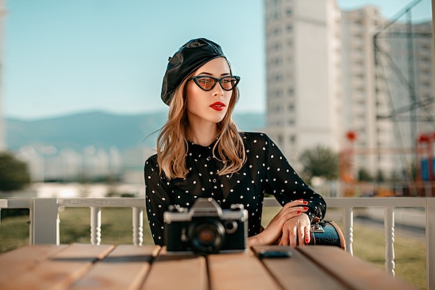 Una mujer joven con un vestido vintage de lunares negros con una vieja cámara en sus manos posando en la calle