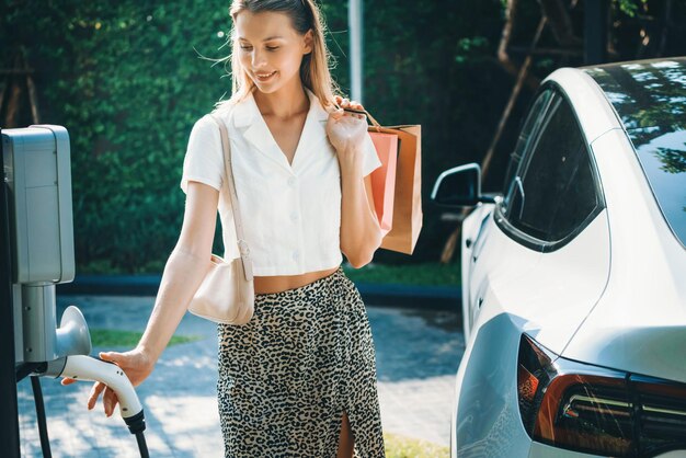 Mujer joven viaja con coche eléctrico eléctrico cargando en el jardín al aire libre de la ciudad sostenible verde en verano Estilo de vida de sostenibilidad urbana por energía limpia y recargable verde de los vehículos eléctricos BEV