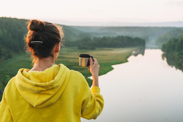 Mujer joven viajera con capucha amarilla con una taza de café de la mañana mirando el amanecer