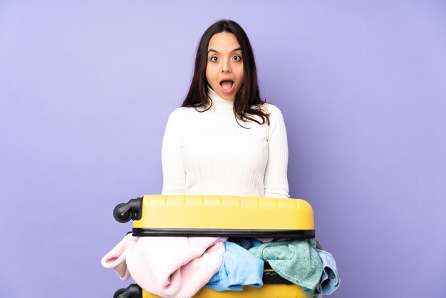 Mujer joven viajero con una maleta llena de ropa sobre pared púrpura aislada con expresión facial sorpresa